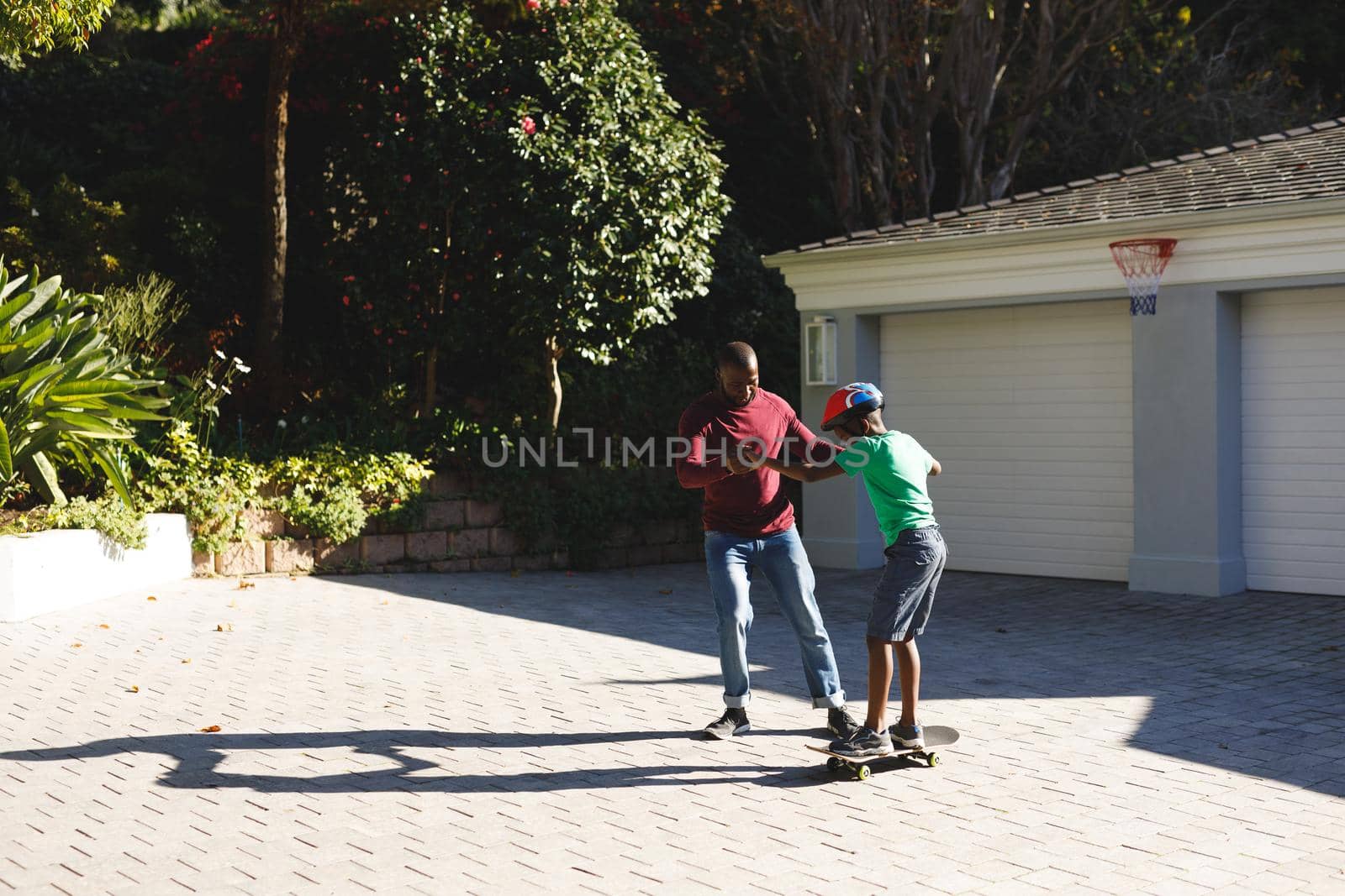 African american father smiling and helping son balancing on skateboard in garden. family spending time at home.