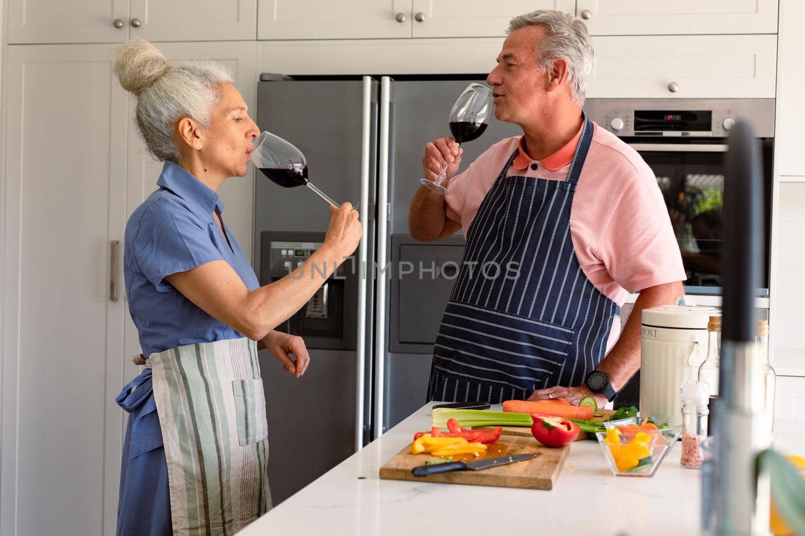 Caucasian senior couple standing in kitchen, drinking wine and preparing meal together. healthy retirement lifestyle at home.