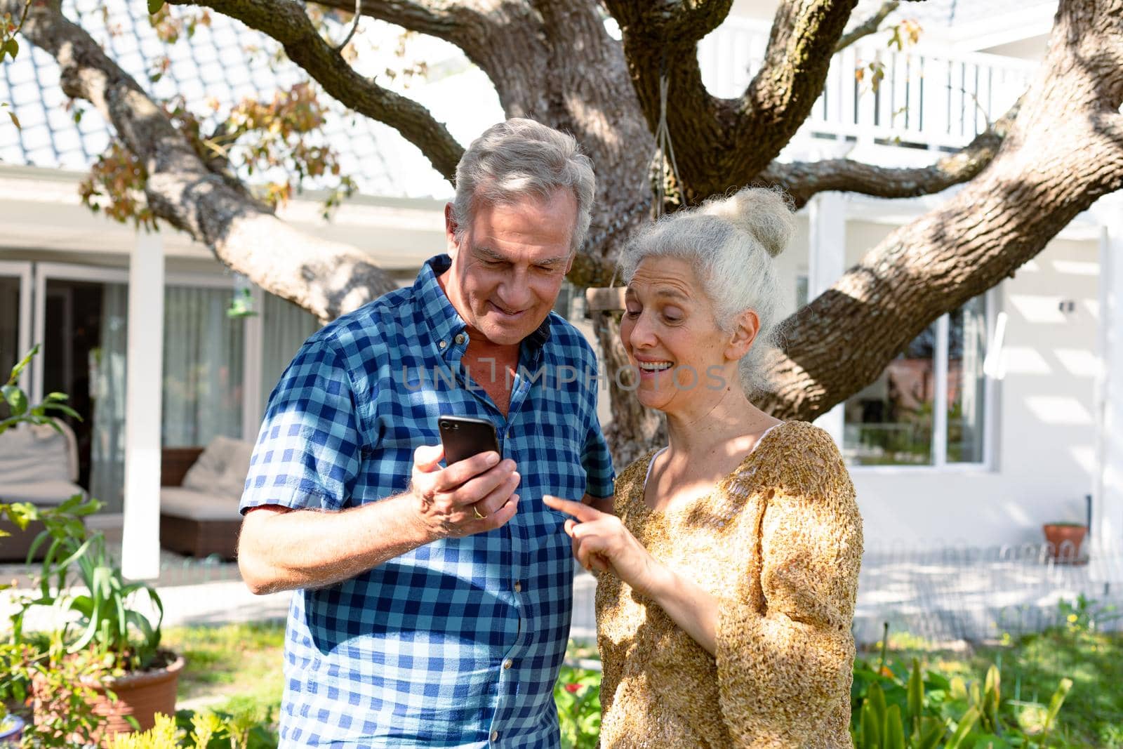 Happy caucasian senior couple using smartphone in front of house. healthy retirement lifestyle at home and garden.