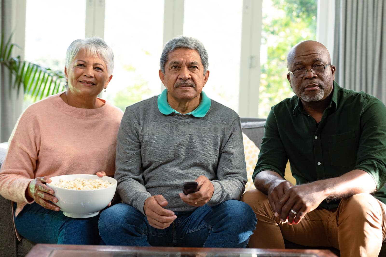 Two focused diverse senior couple and their african american male friend watching tv and having fun. retirement lifestyle relaxing at home with technology.
