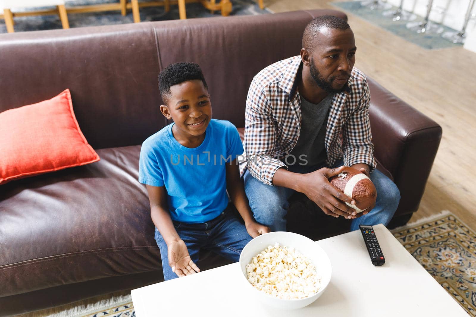 Happy african american father and son sitting on sofa, watching match on tv. family spending time together at home.