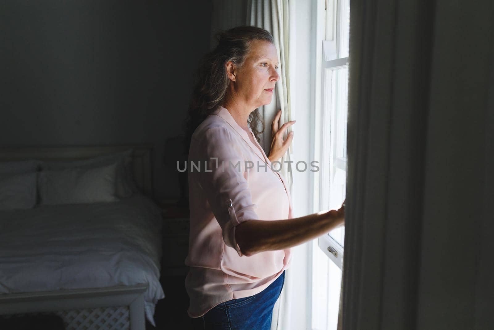 Thoughtful senior caucasian woman in bedroom, standing next to window, widening curtains. retirement lifestyle, spending time alone at home.