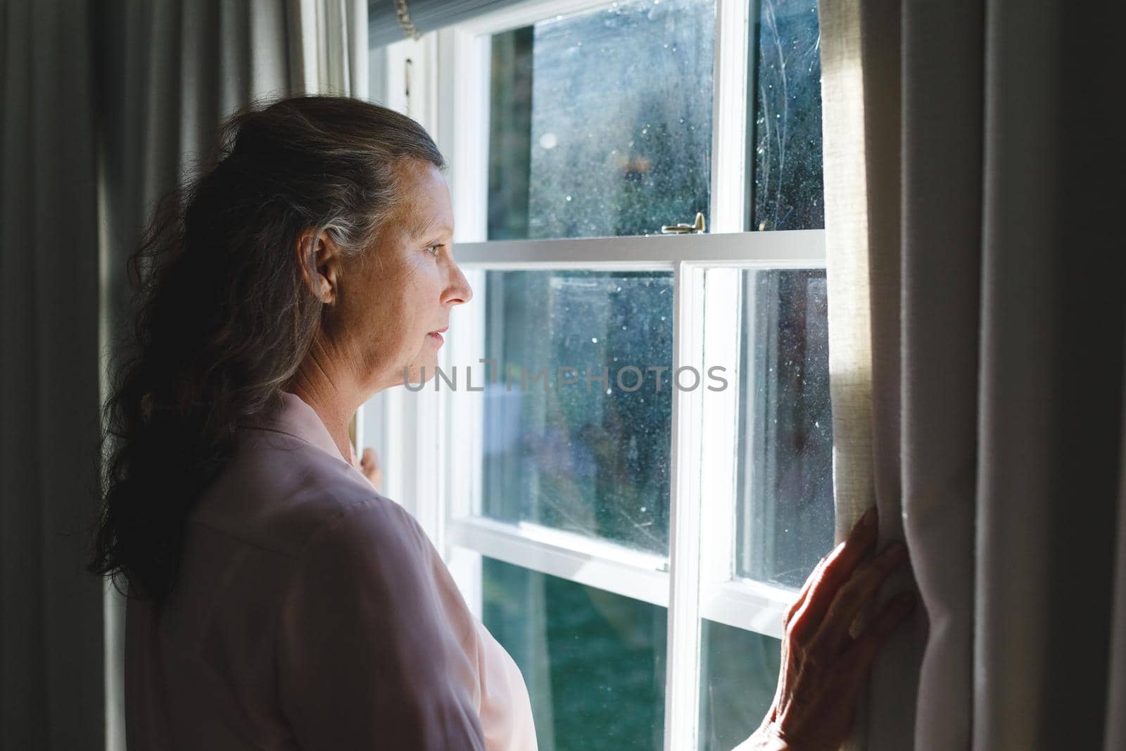 Thoughtful senior caucasian woman in bedroom, standing next to window, widening curtains. retirement lifestyle, spending time alone at home.