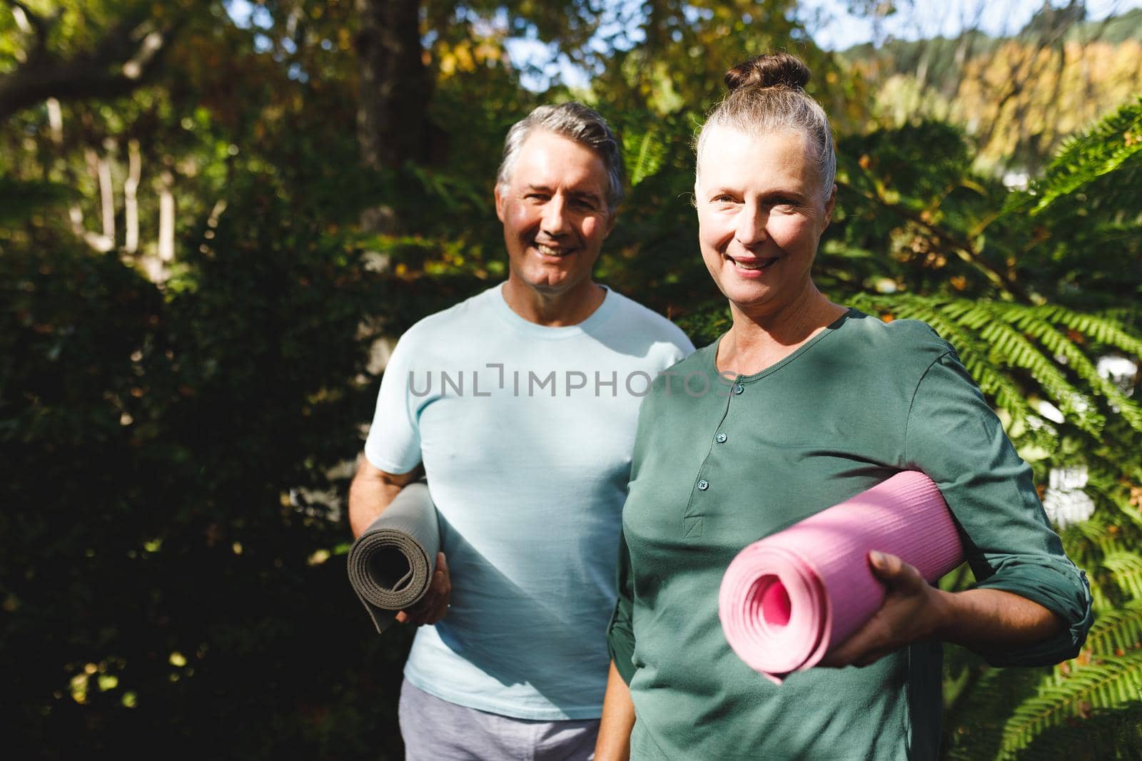 Portrait of happy senior caucasian couple holding yoga mats, looking to camera in sunny garden. healthy retirement lifestyle, spending time at home.