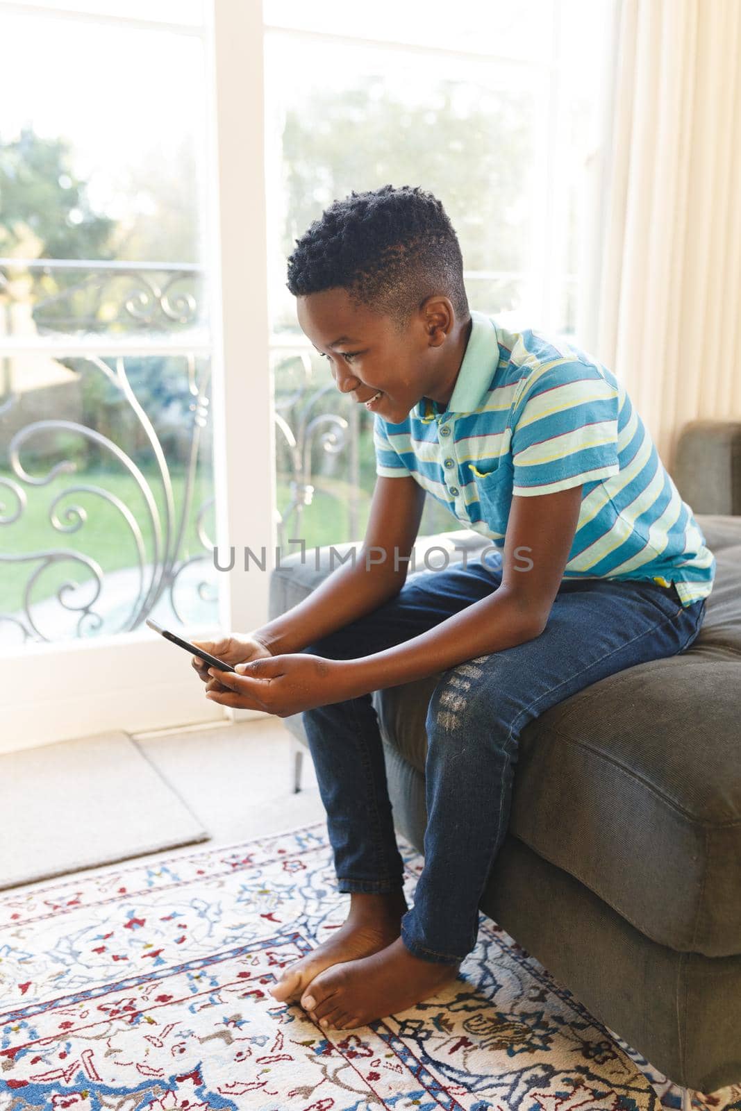 Happy african american boy using smartphone and sitting on couch in living room. spending time alone with technology at home.
