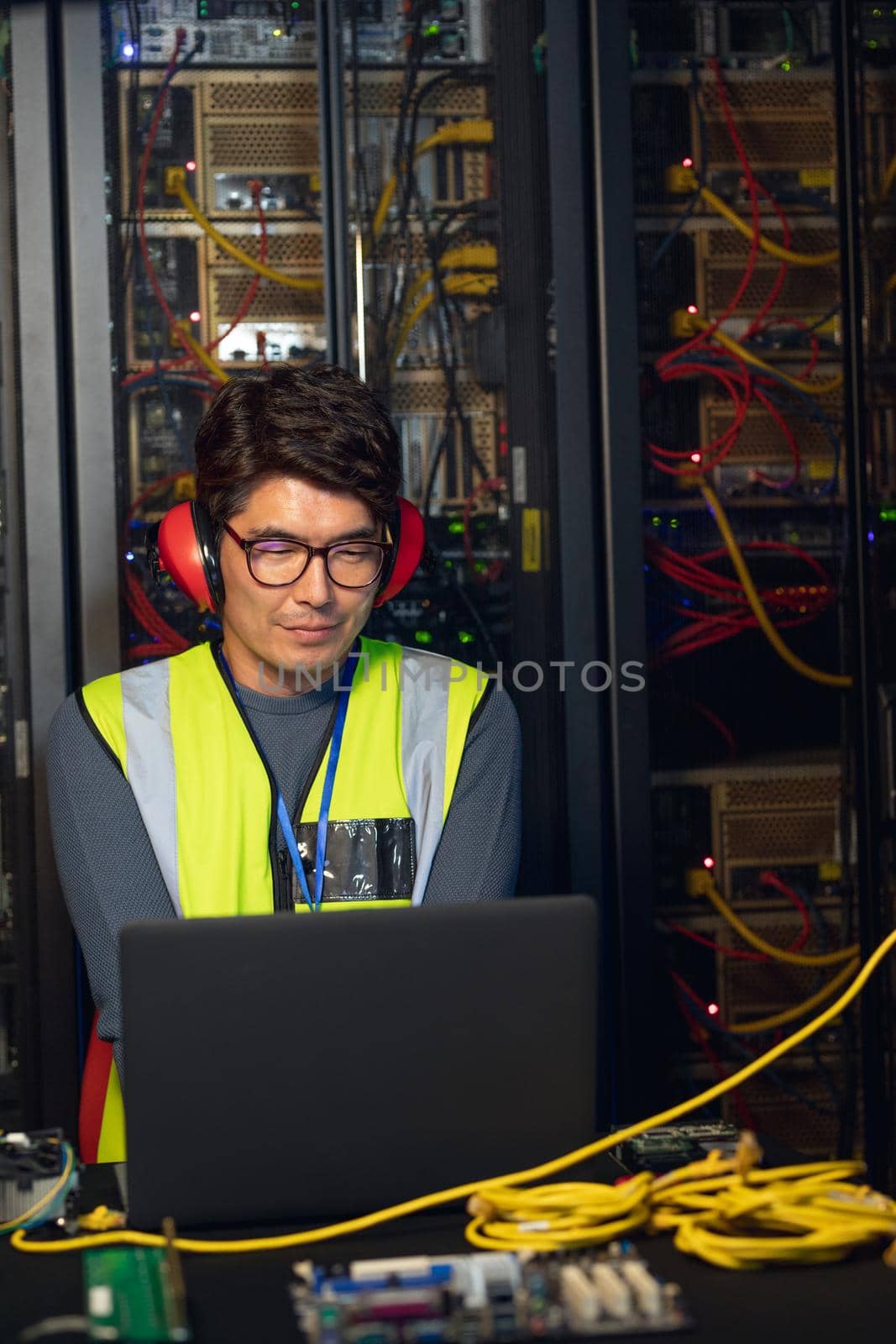 Asian male engineer wearing ear plugs using a laptop in computer server room. database server management and maintenance concept