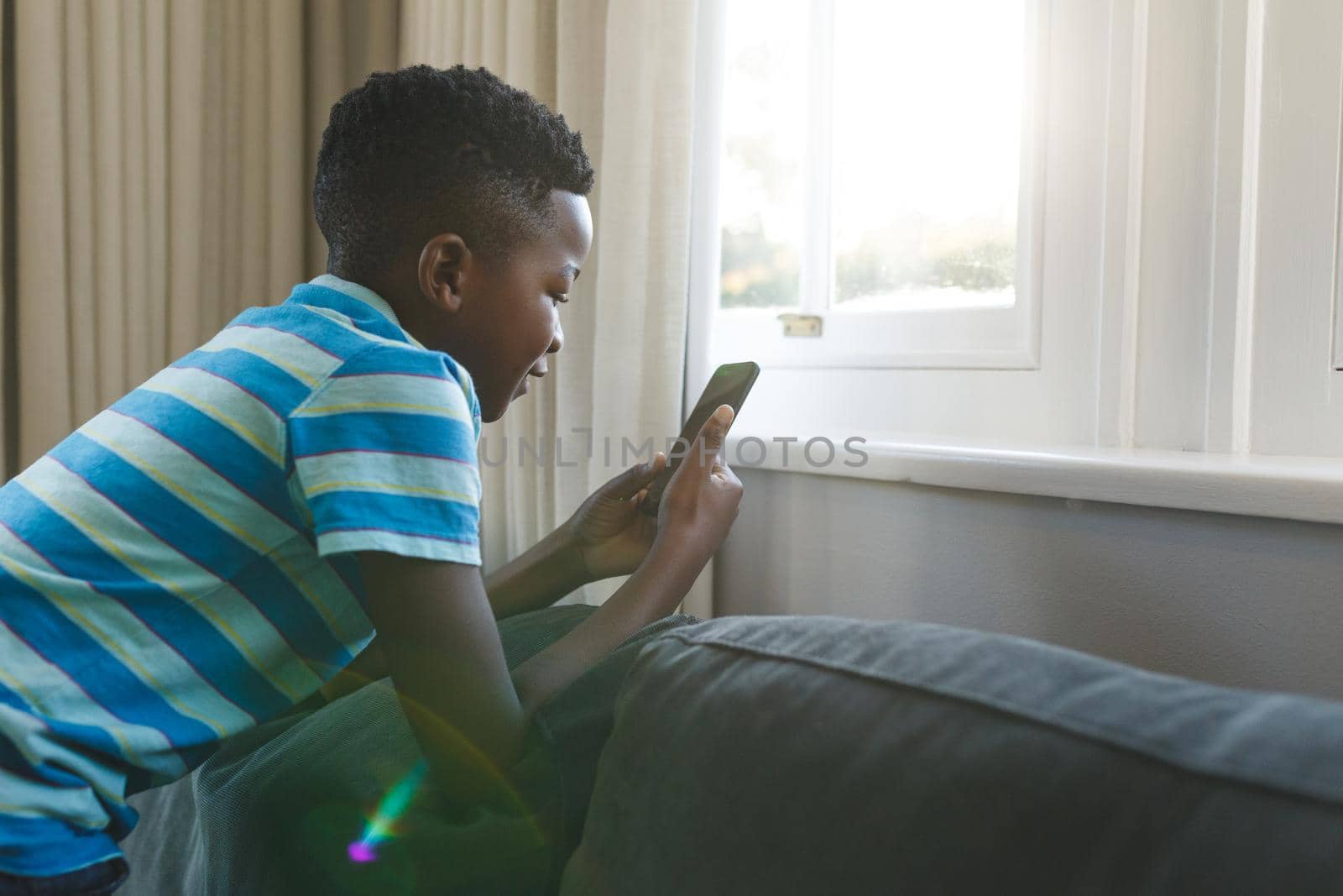 Happy african american boy kneeling on couch using smartphone in living room. spending time alone at home with technology.