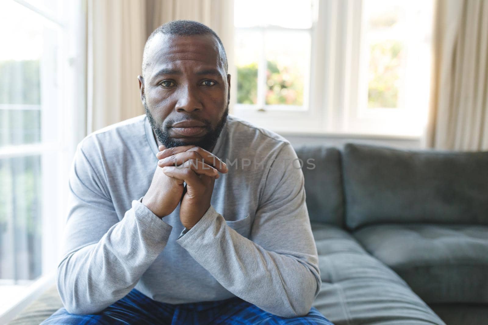 Portrait of sad african american man thinking and sitting with chin on hands in living room. spending time alone at home.