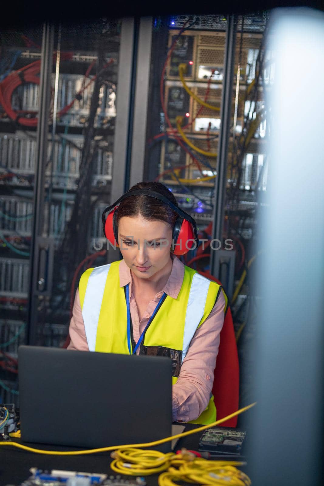Caucasian female engineer wearing ear plugs using a laptop in computer server room. database server management and maintenance concept