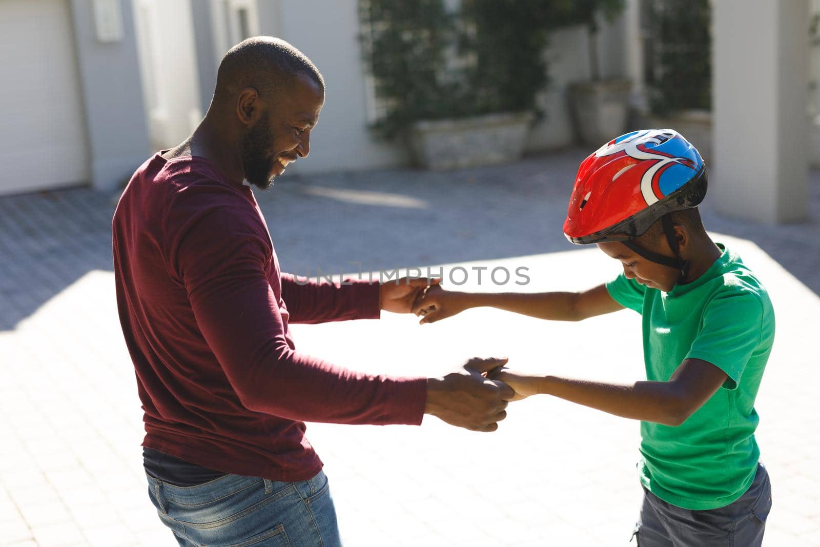 African american father smiling and helping son balancing on skateboard in garden by Wavebreakmedia