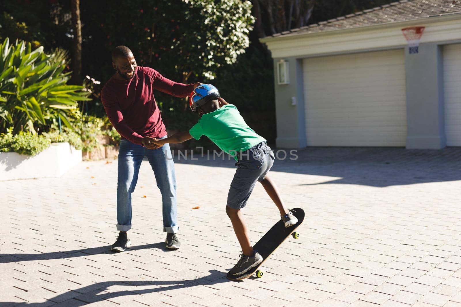 African american father smiling and helping son balancing on skateboard in garden. family spending time at home.
