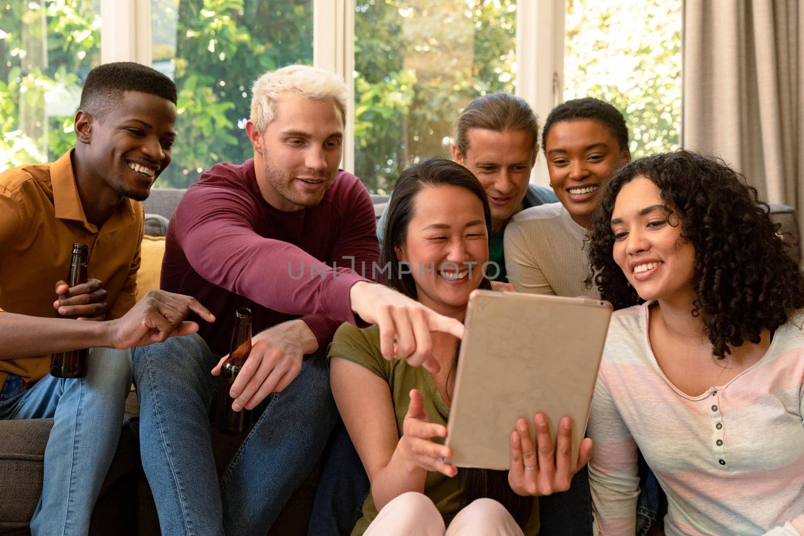 Group of happy diverse female and male friends drinking beer together and using tablet. socialising with friends at home.