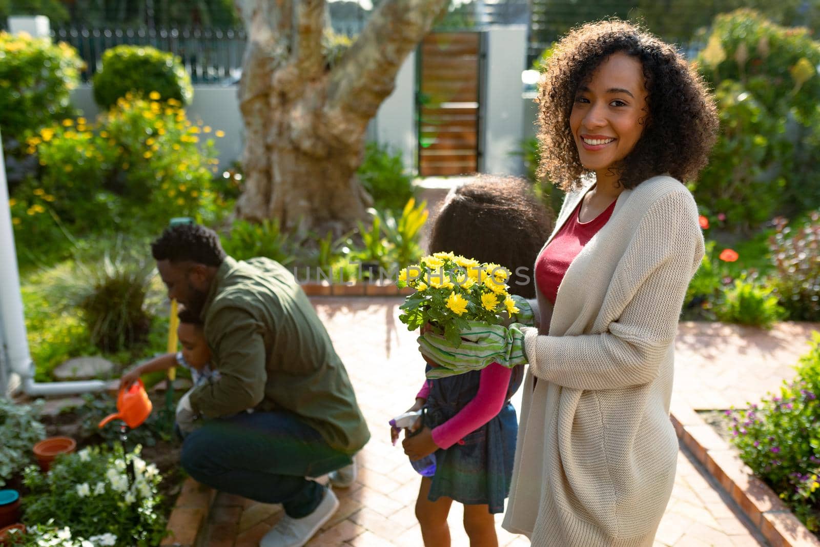 Happy african american mother holding flowers, gardening with children and husband. family time, having fun together at home and garden.