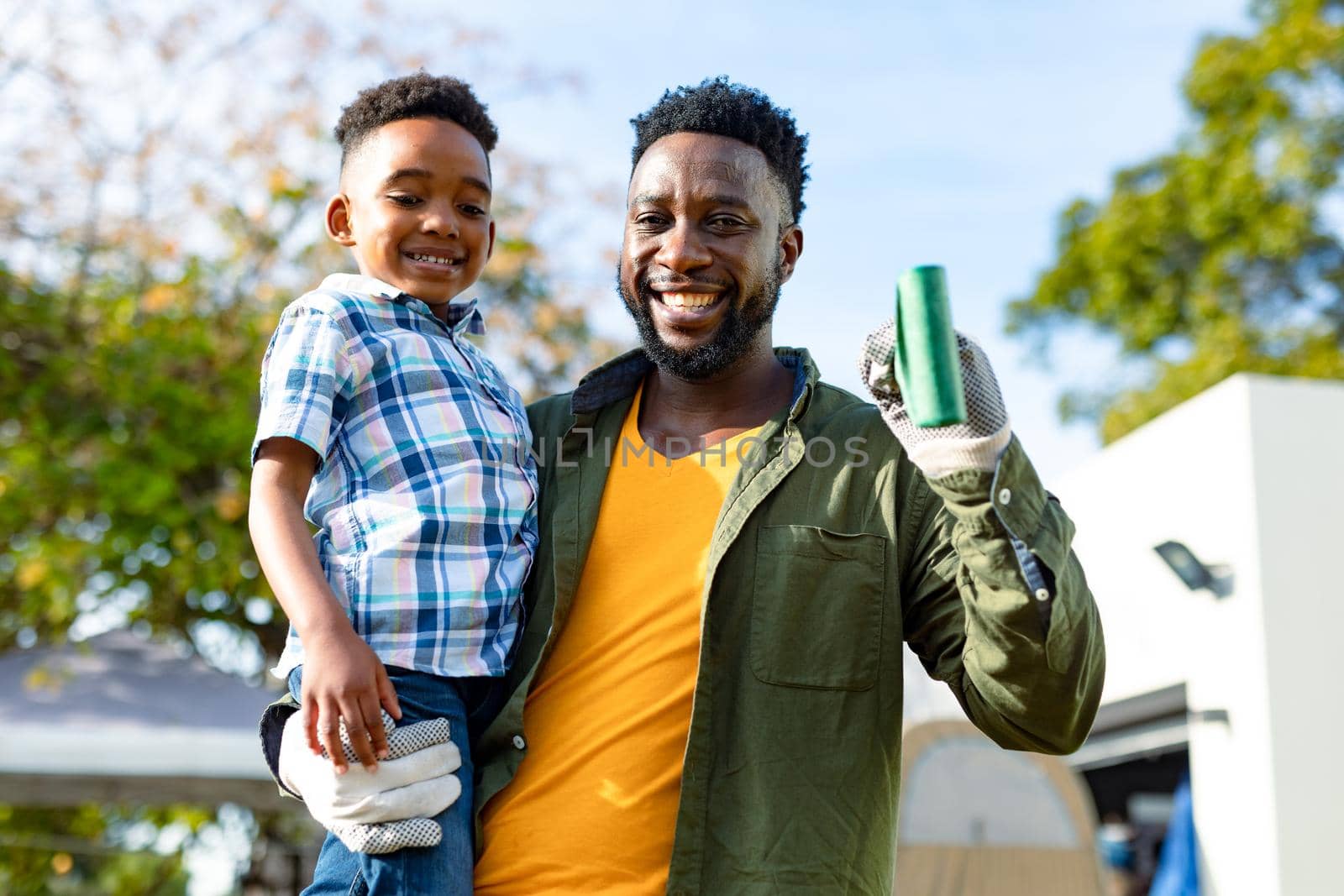 Happy african american father holding his son and pitchfork. family time, having fun together at home and garden.