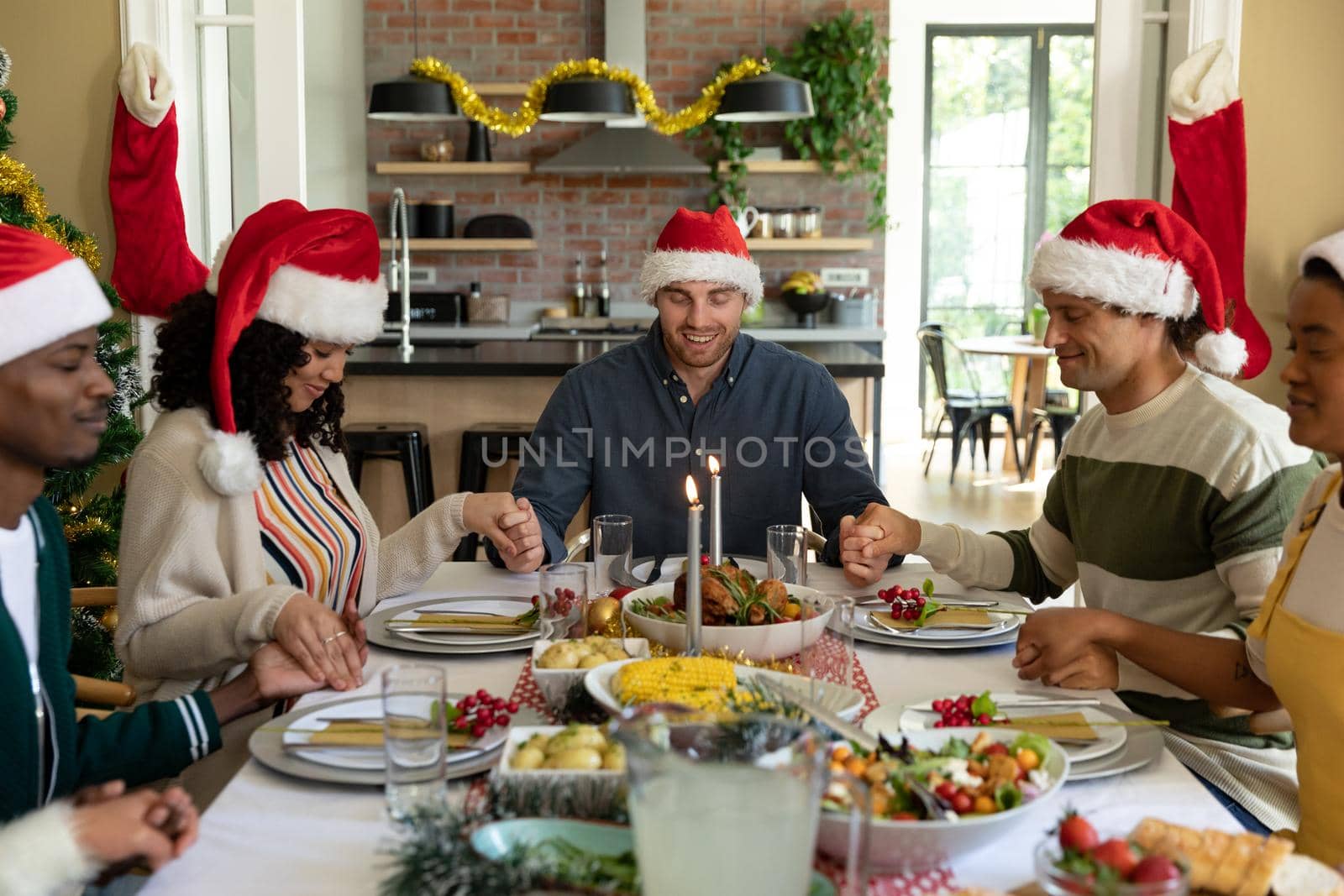 Group of happy diverse female and male friends having christmas meal, praying together at home. christmas festivities, celebrating at home with friends
