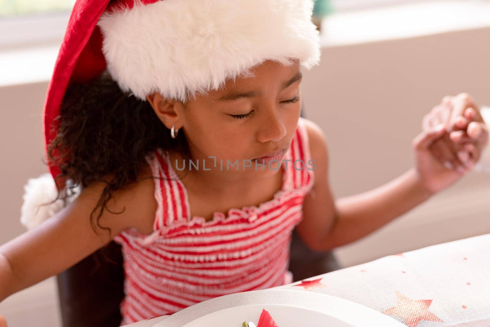 African american girl wearing santa hat praying at christmas table. christmas, festivity and tradition at home.