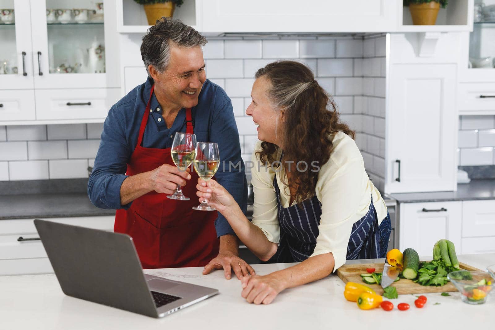 Happy senior caucasian couple in kitchen cooking together, drinking wine, using laptop. retirement lifestyle, spending time at home.