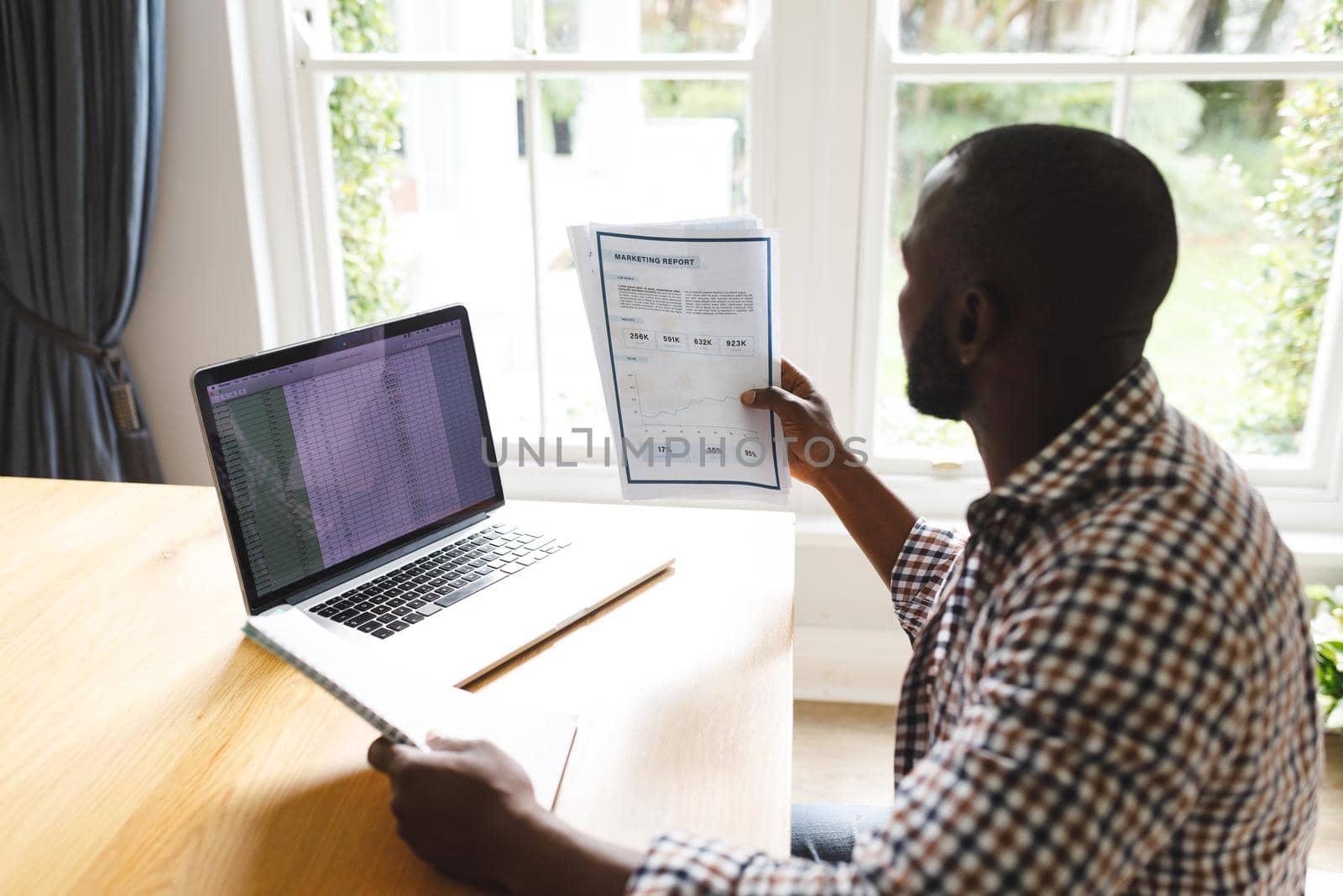 African american man sitting at table with paperwork in dining room, working remotely using laptop by Wavebreakmedia