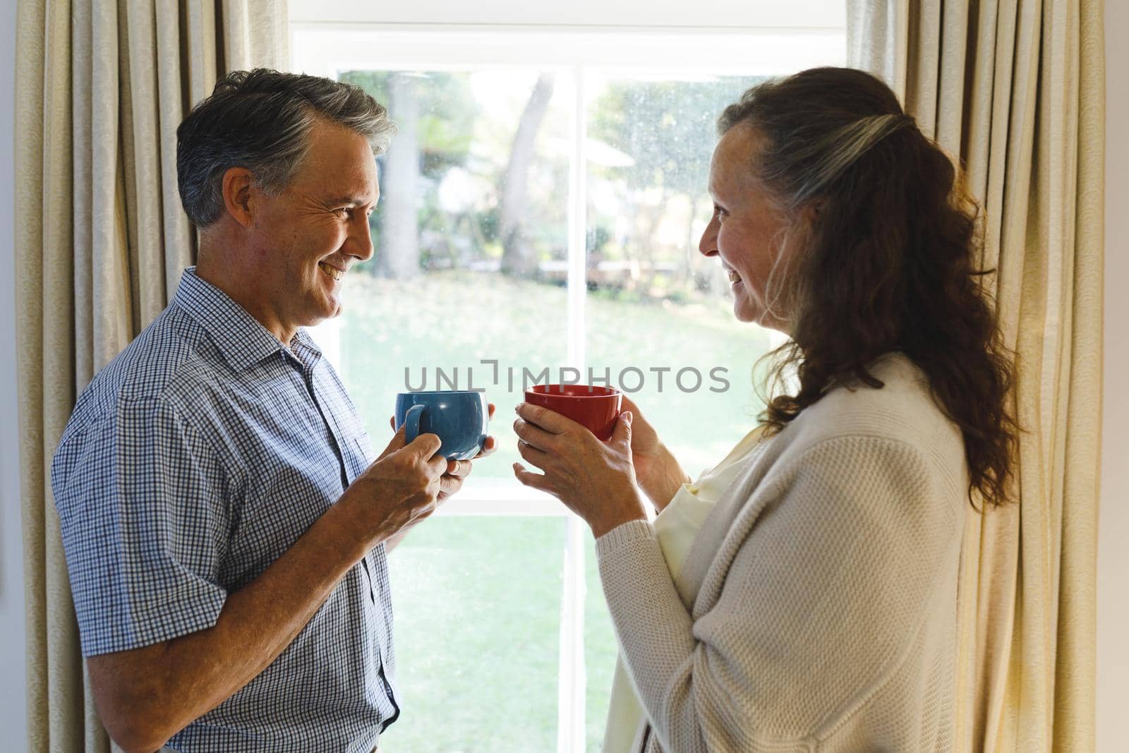 Happy senior caucasian couple standing next to window, holding cups. retirement lifestyle, spending time at home.