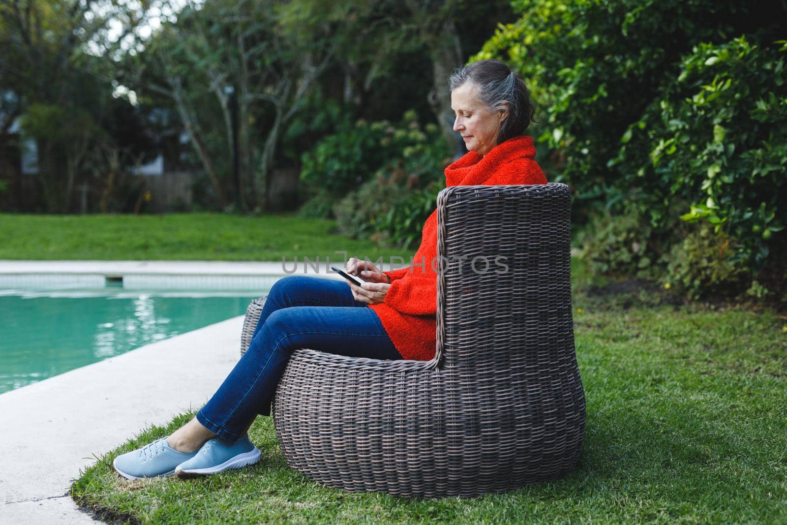 Happy senior caucasian woman sitting on chair, using smartphone in garden. retirement lifestyle, spending time alone at home with technology.