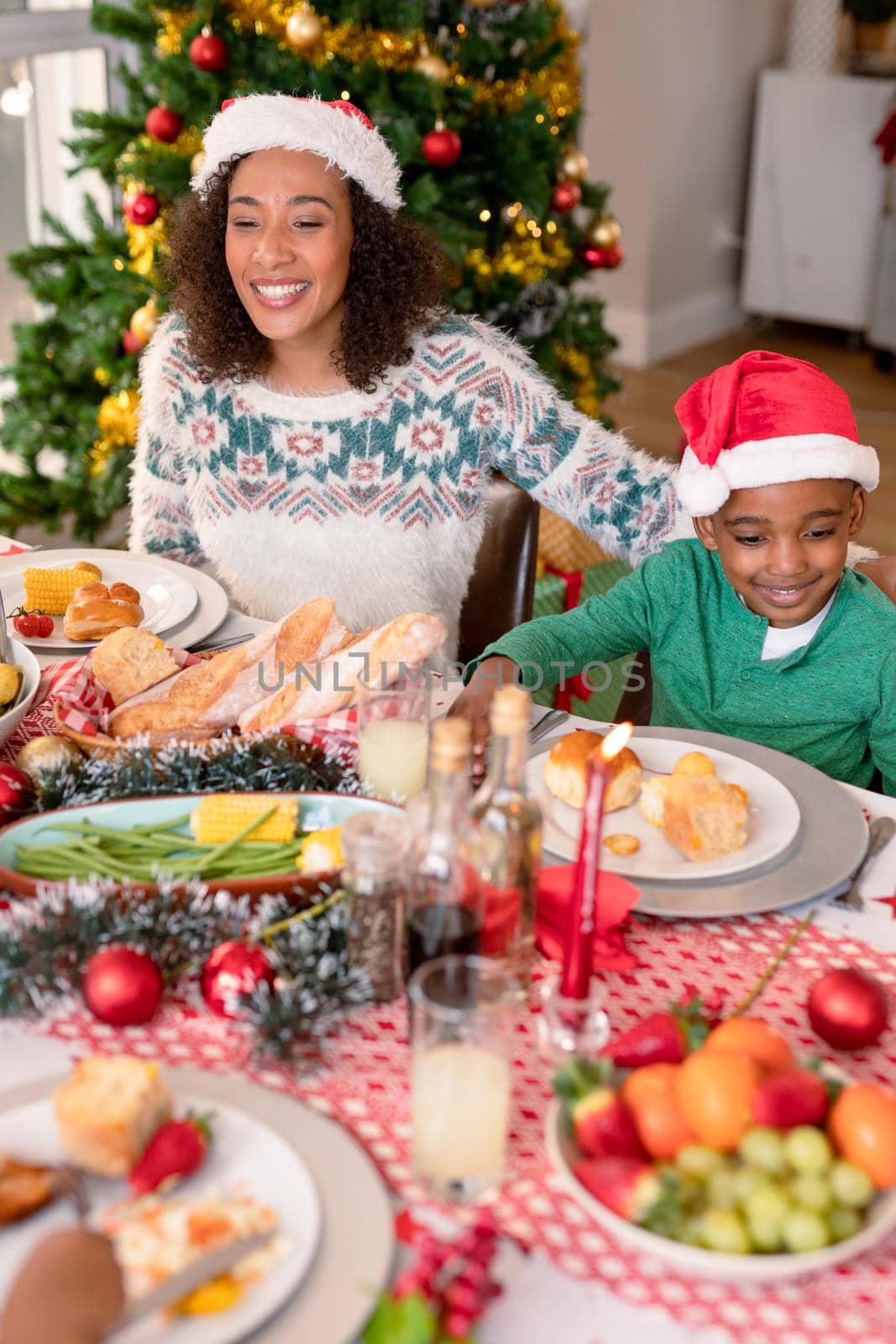 Happy african american mother and son wearing santa hats sitting at christmas table. family christmas time and festivity together at home.