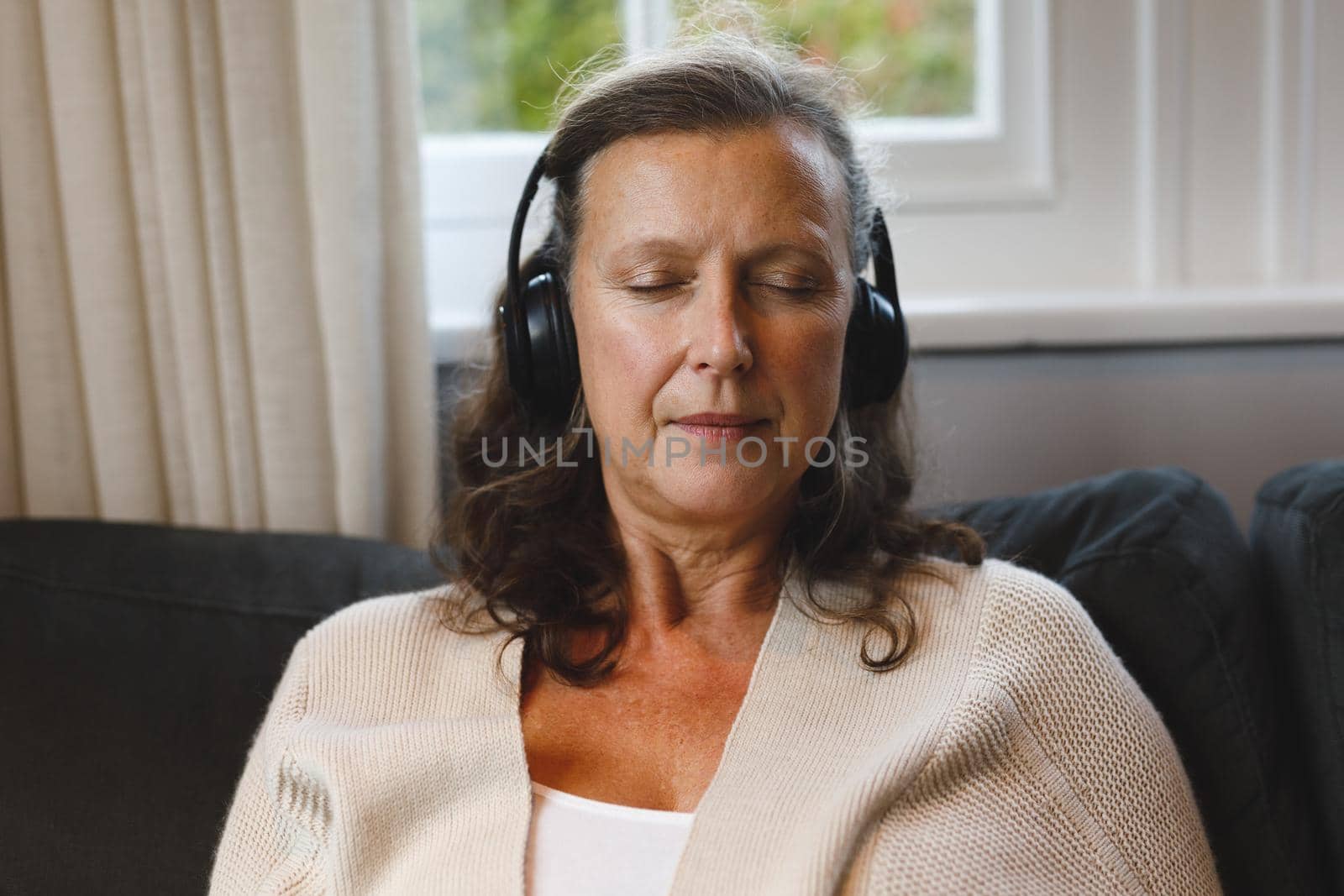 Happy senior caucasian woman in living room sitting on sofa, wearing headphones. retirement lifestyle, spending time alone at home with technology.