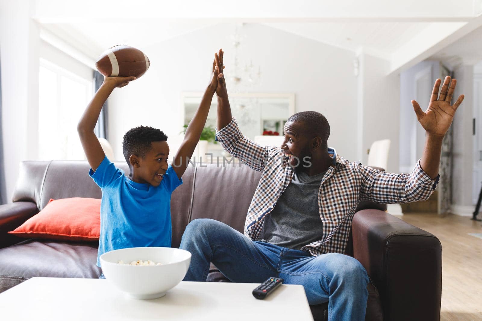 Excited african american father and son sitting on sofa, watching match on tv and cheering. family spending time together at home.