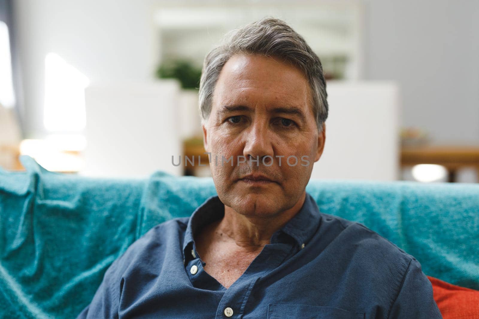 Portrait of sad senior caucasian man in living room sitting on sofa, looking to camera. retirement lifestyle, spending time at home.