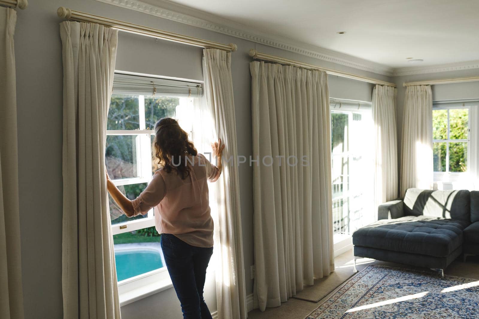 Thoughtful senior caucasian woman in bedroom, standing next to window, widening curtains. retirement lifestyle, spending time alone at home.