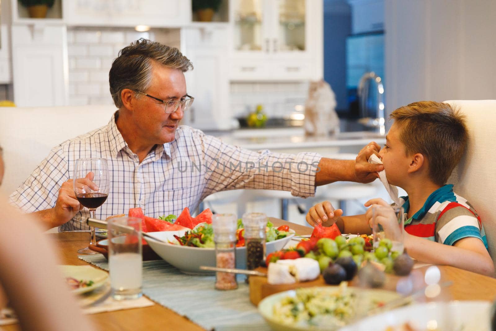 Caucasian grandfather wiping face of grandson while sitting at table and having dinner by Wavebreakmedia