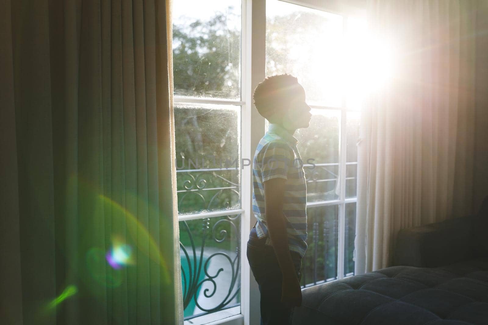 African american boy thinking and standing at window in sunny living room. spending time alone at home.