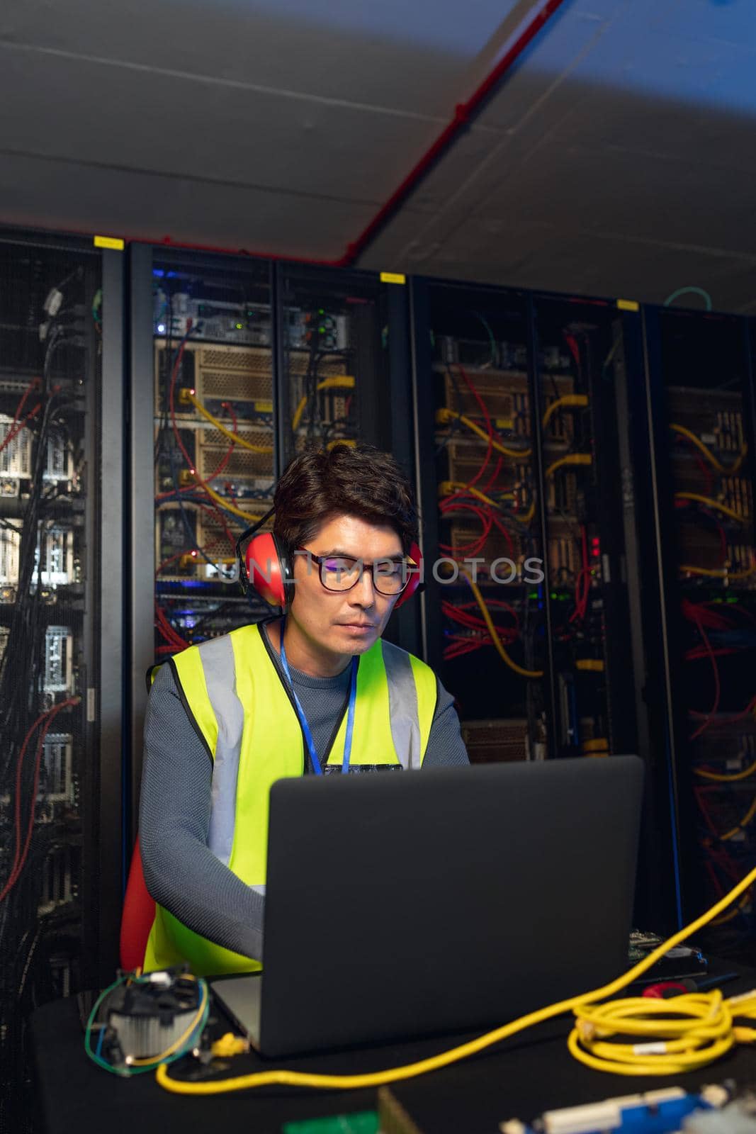 Asian male engineer wearing ear plugs using a laptop in computer server room by Wavebreakmedia