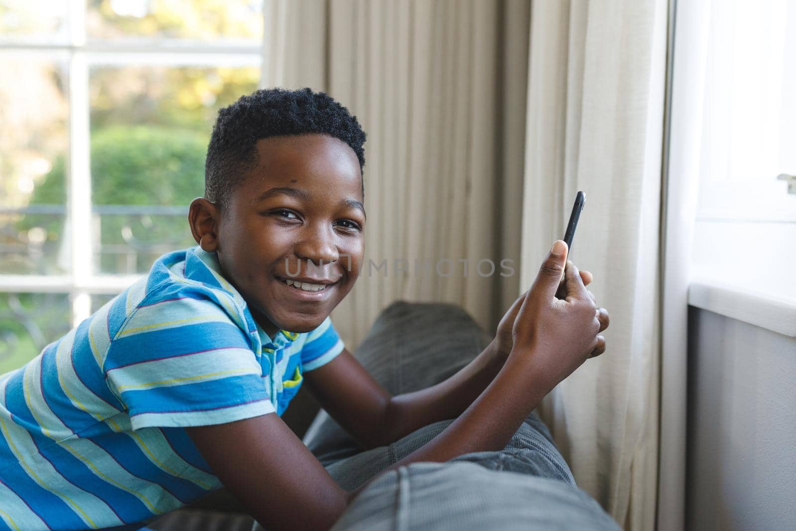 Portrait of happy african american boy kneeling on couch using smartphone in living room by Wavebreakmedia