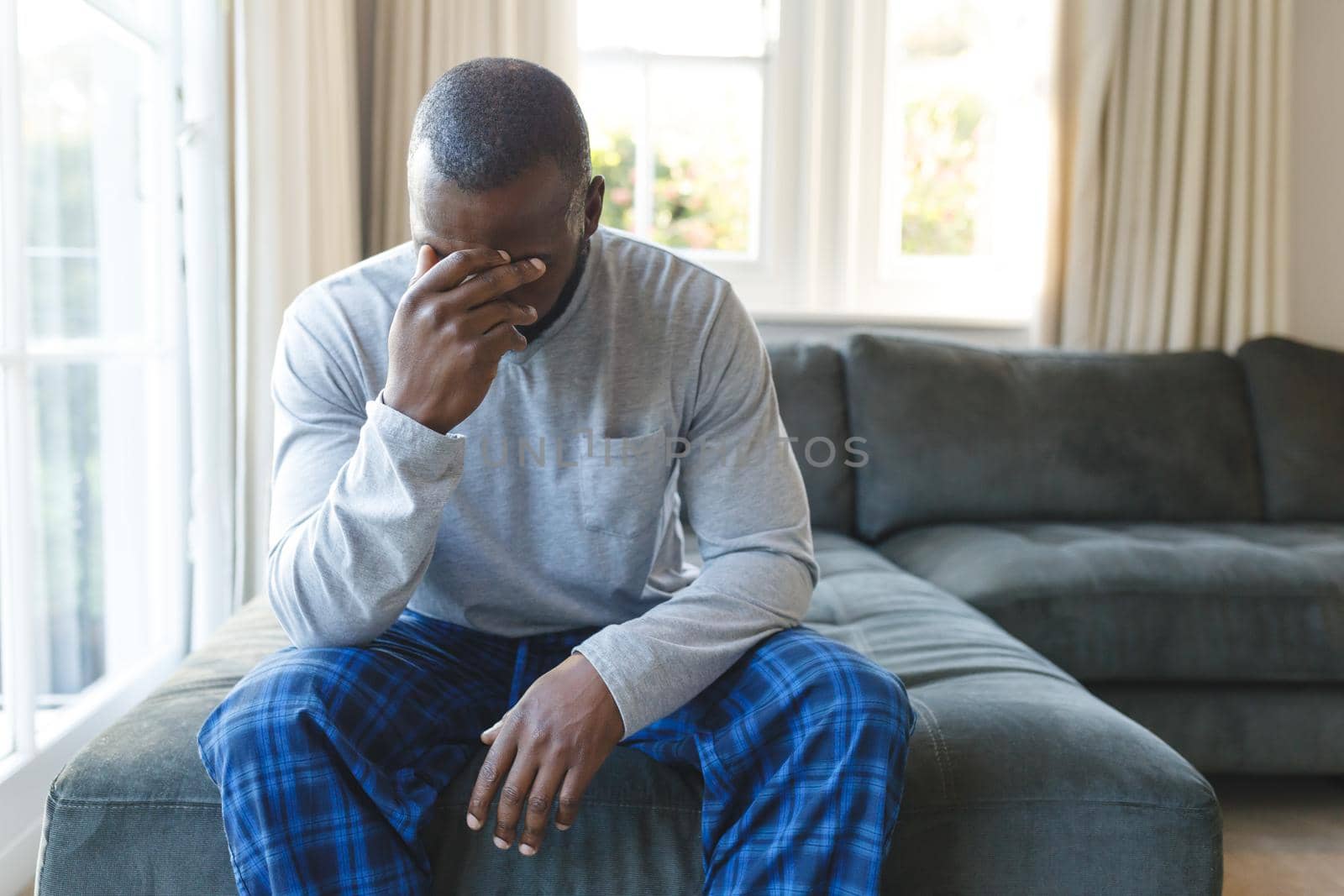 Sad african american man covering his face sitting on couch by window in living room. spending time alone at home.