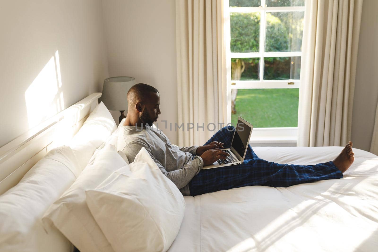 African american man using laptop and lying on bed in his bedroom. spending time alone at home with technology.