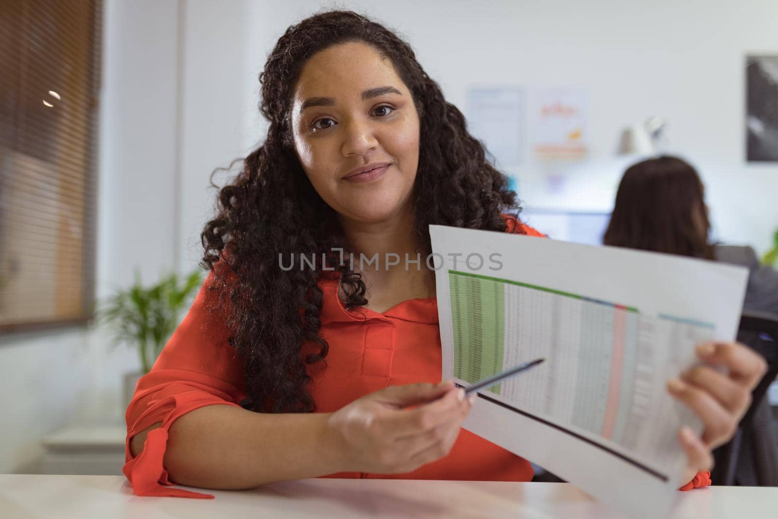 Smiling biracial businesswoman sitting at desk making video call holding documents in modern office. business and office workplace.
