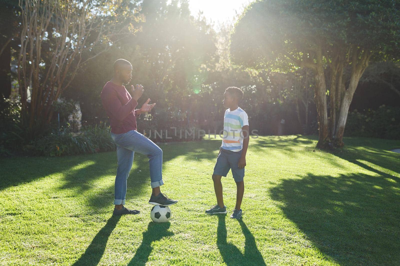 African american father talking with son and playing football in garden. family spending time at home.
