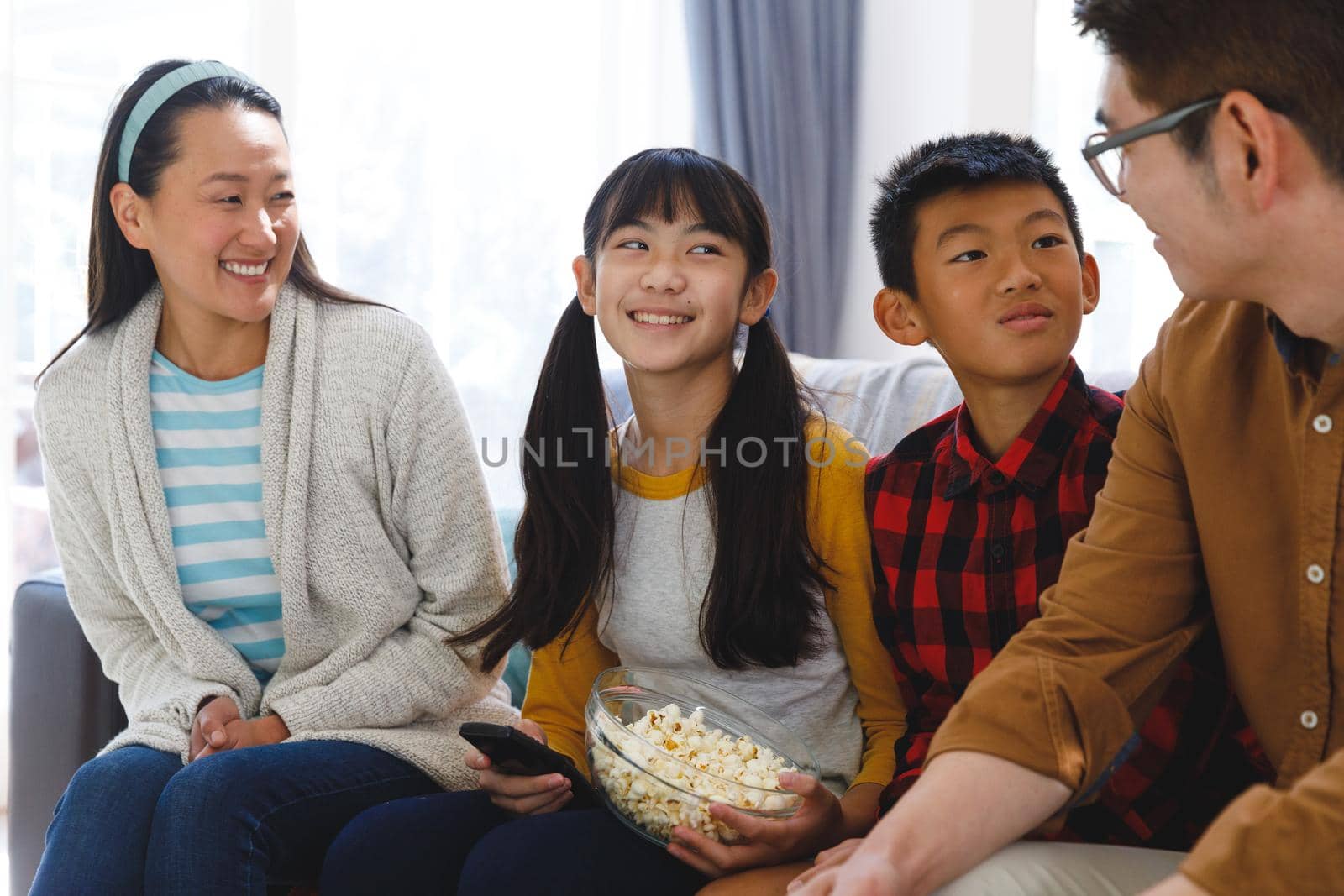 Smiling asian parents with son and daughter sitting on couch and watching tv with popcorn. family entertainment and leisure time together at home.