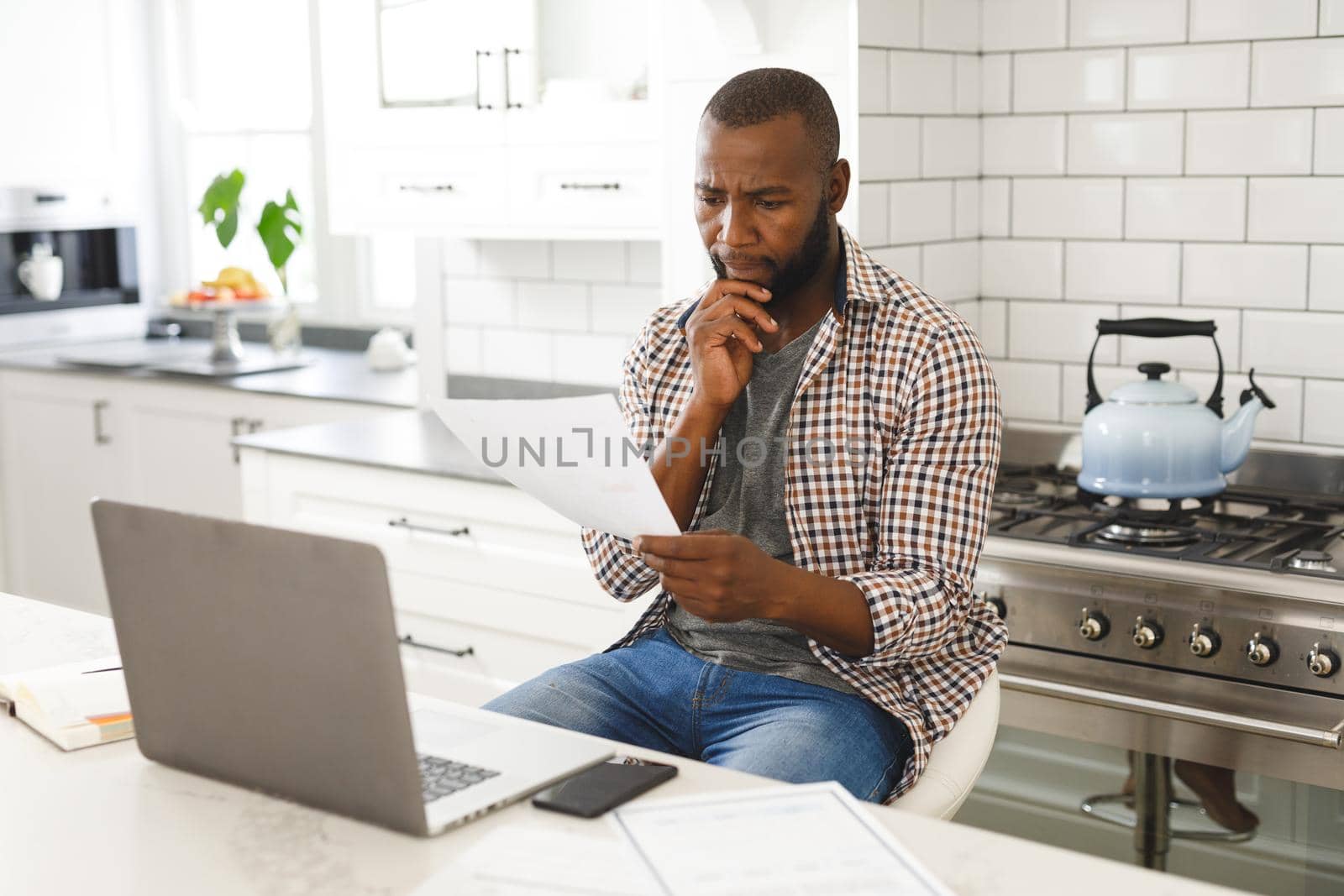 Serious african american man sitting in kitchen working looking at paperwork and using laptop. remote working from home with technology.