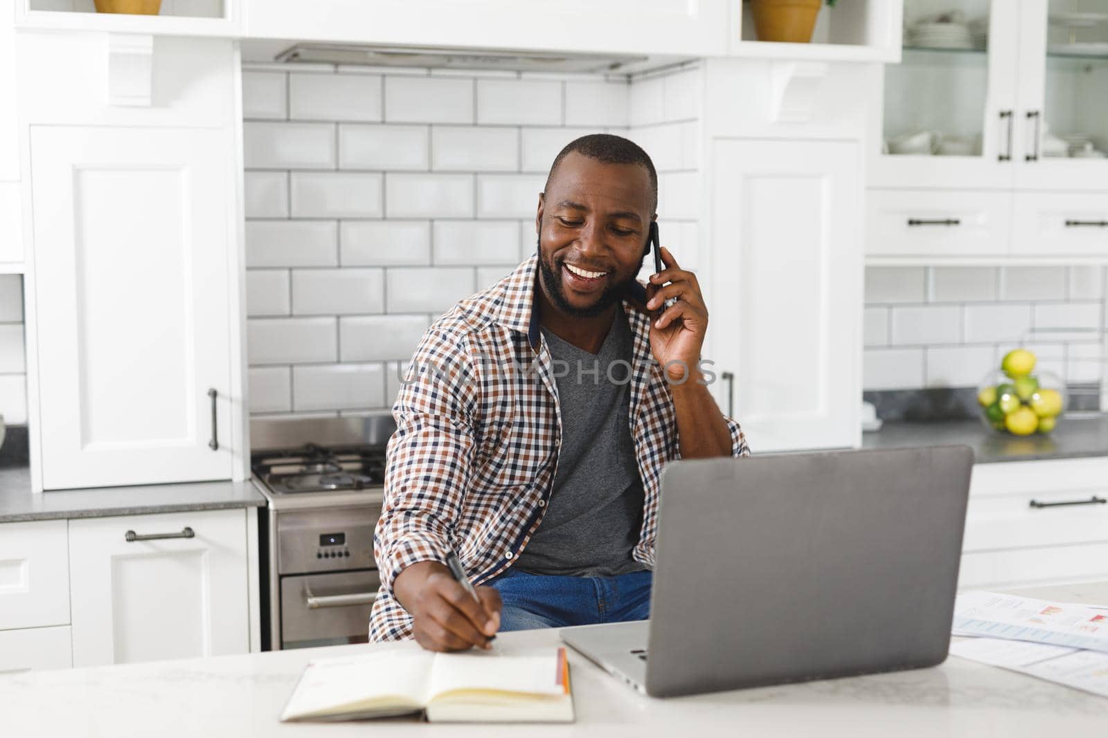 Smiling african american man sitting in kitchen working using laptop and smartphone. remote working from home with technology.