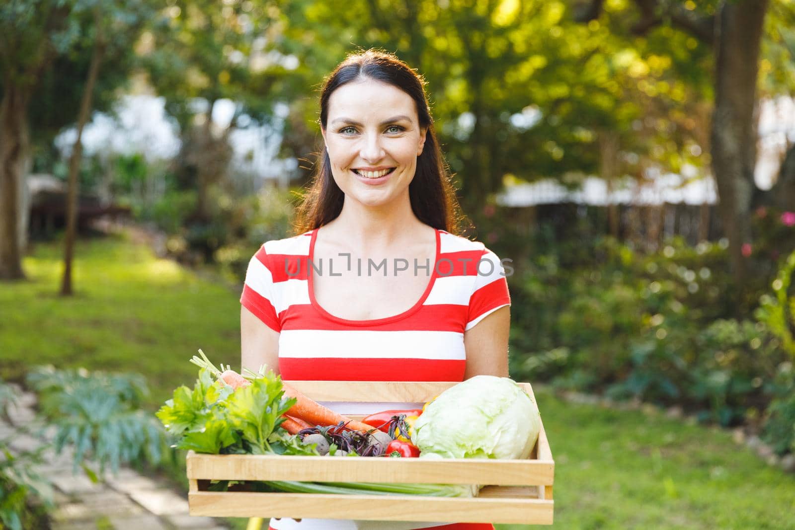 Portrait of smiling caucasian woman standing in garden holding box of fresh organic vegetables. gardening, self sufficiency and growing home produce.