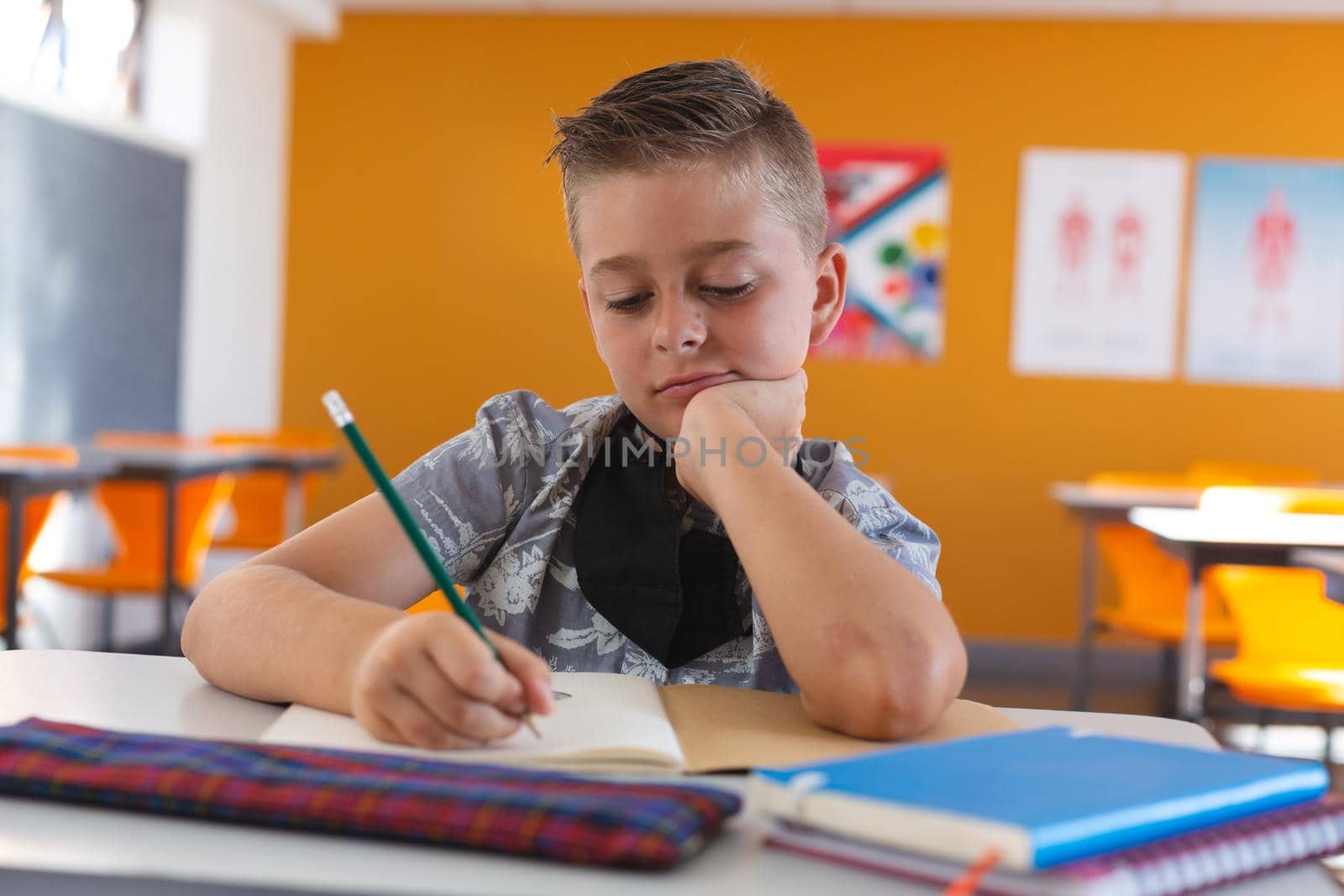 Caucasian schoolboy with face mask sitting in classroom concentrating and writing in schoolbook. childhood and education at elementary school during coronavirus covid19 pandemic.