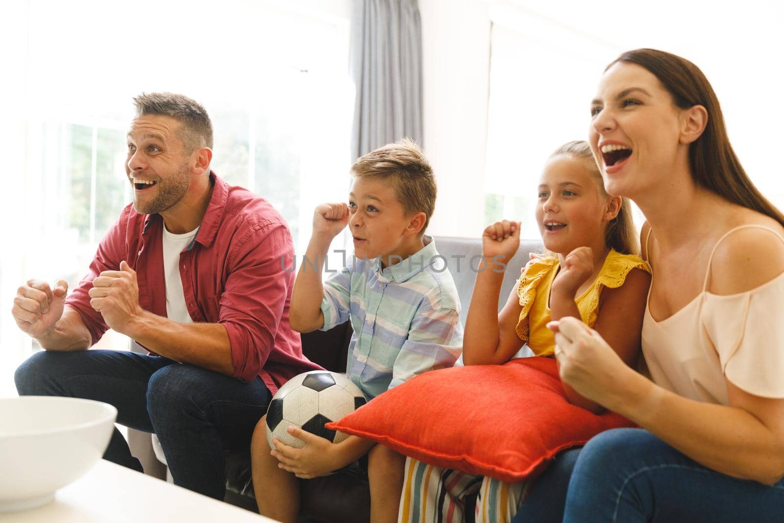 Excited caucasian parents on couch with daughter and son watching football match on tv and cheering by Wavebreakmedia