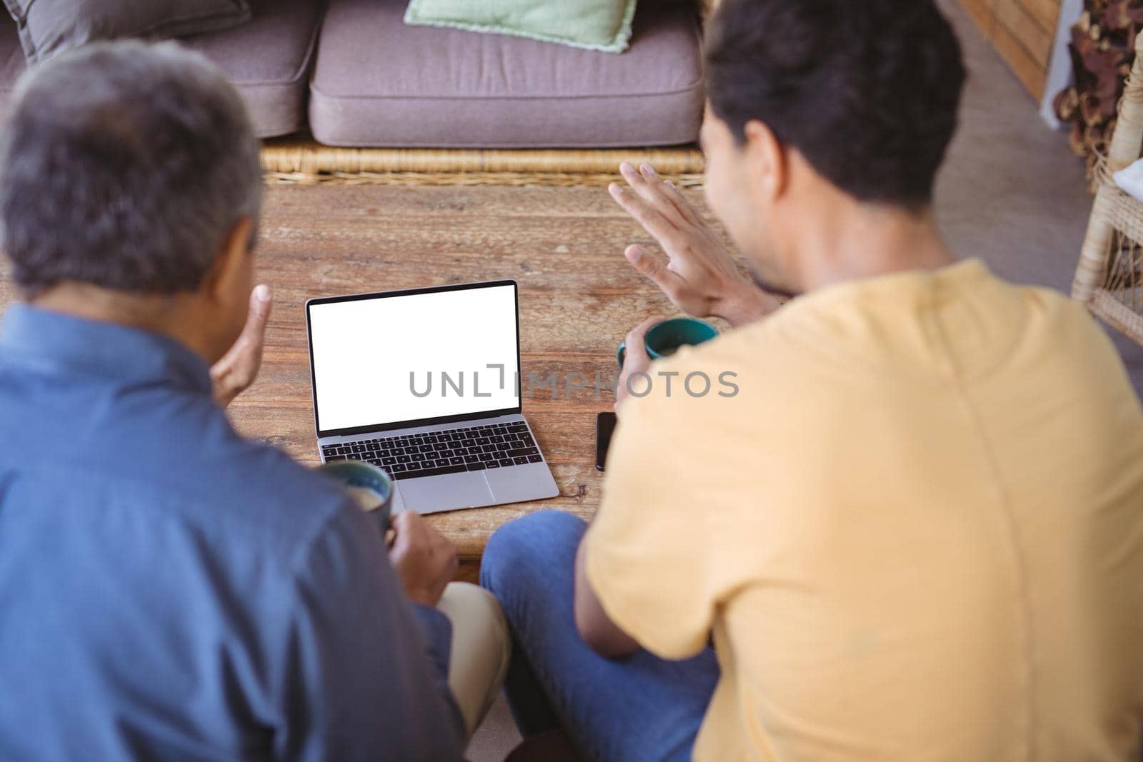 Biracial adult son and senior father holding cups of coffee and making laptop video call. family time at home using technology together.