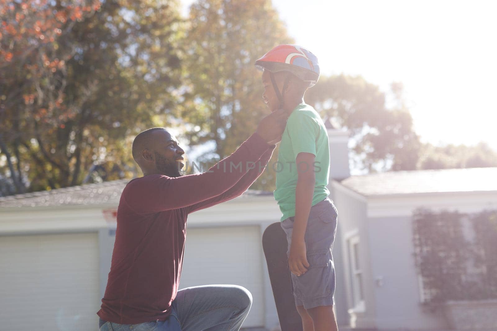 African american father with son smiling and preparing before skateboarding in sunny garden. family spending time at home.