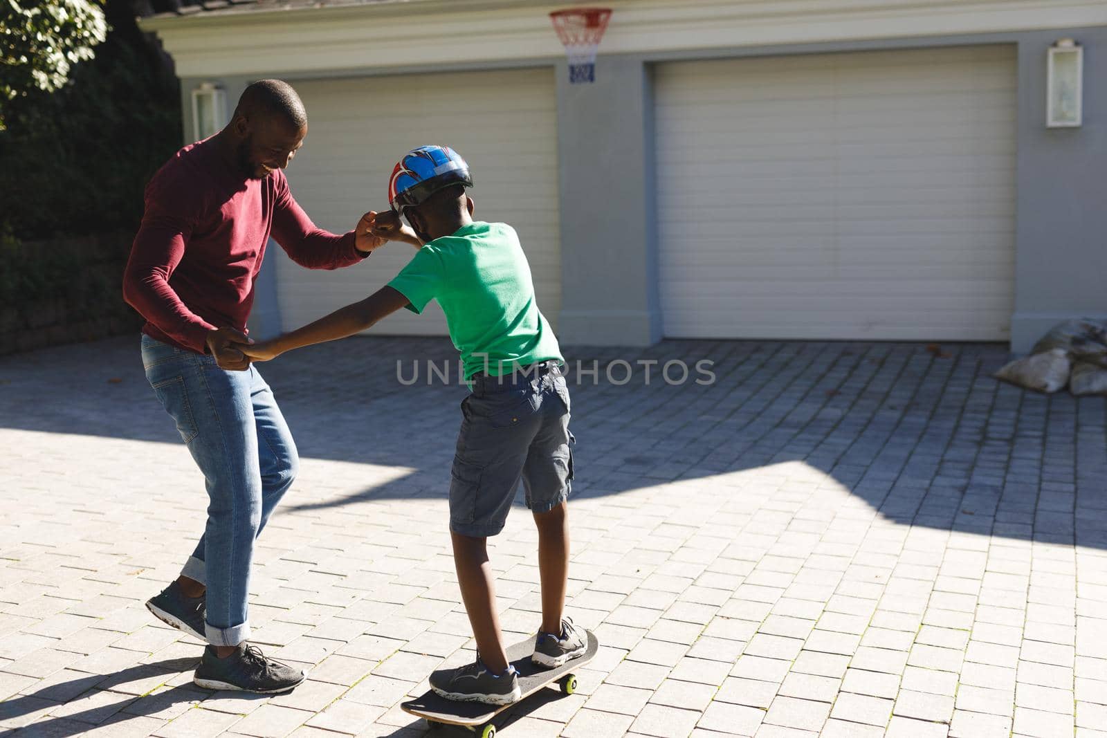 African american father smiling and helping son balancing on skateboard in garden by Wavebreakmedia