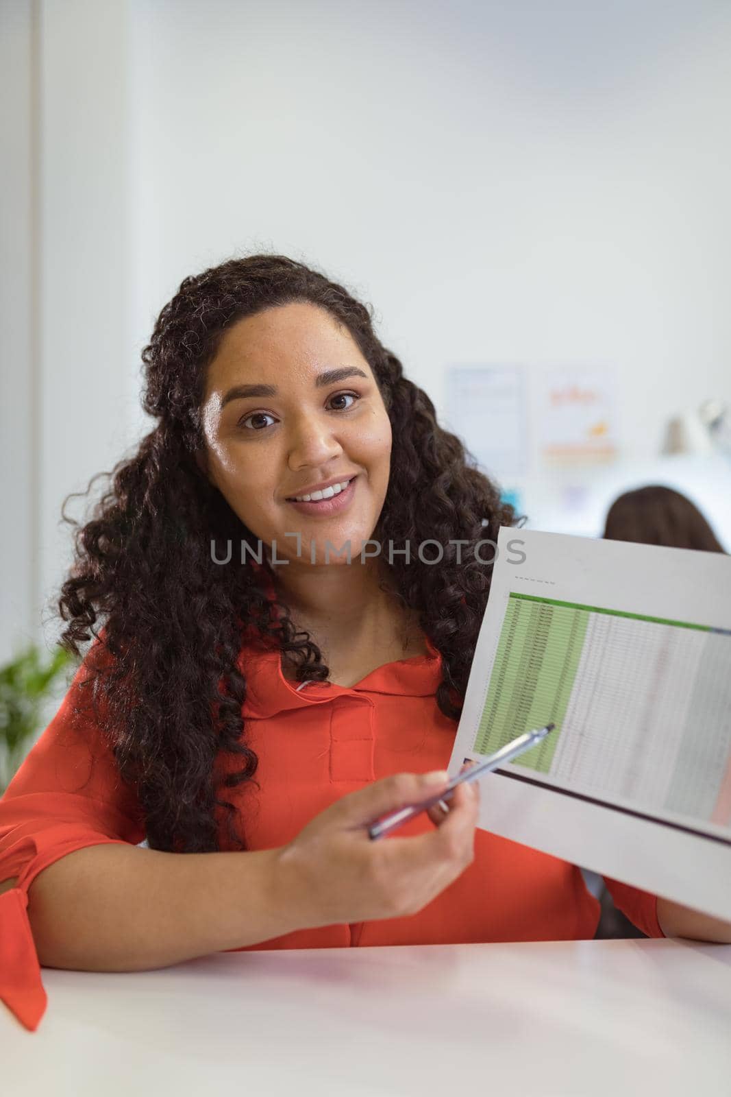Smiling biracial businesswoman sitting at desk making video call holding documents in modern office by Wavebreakmedia
