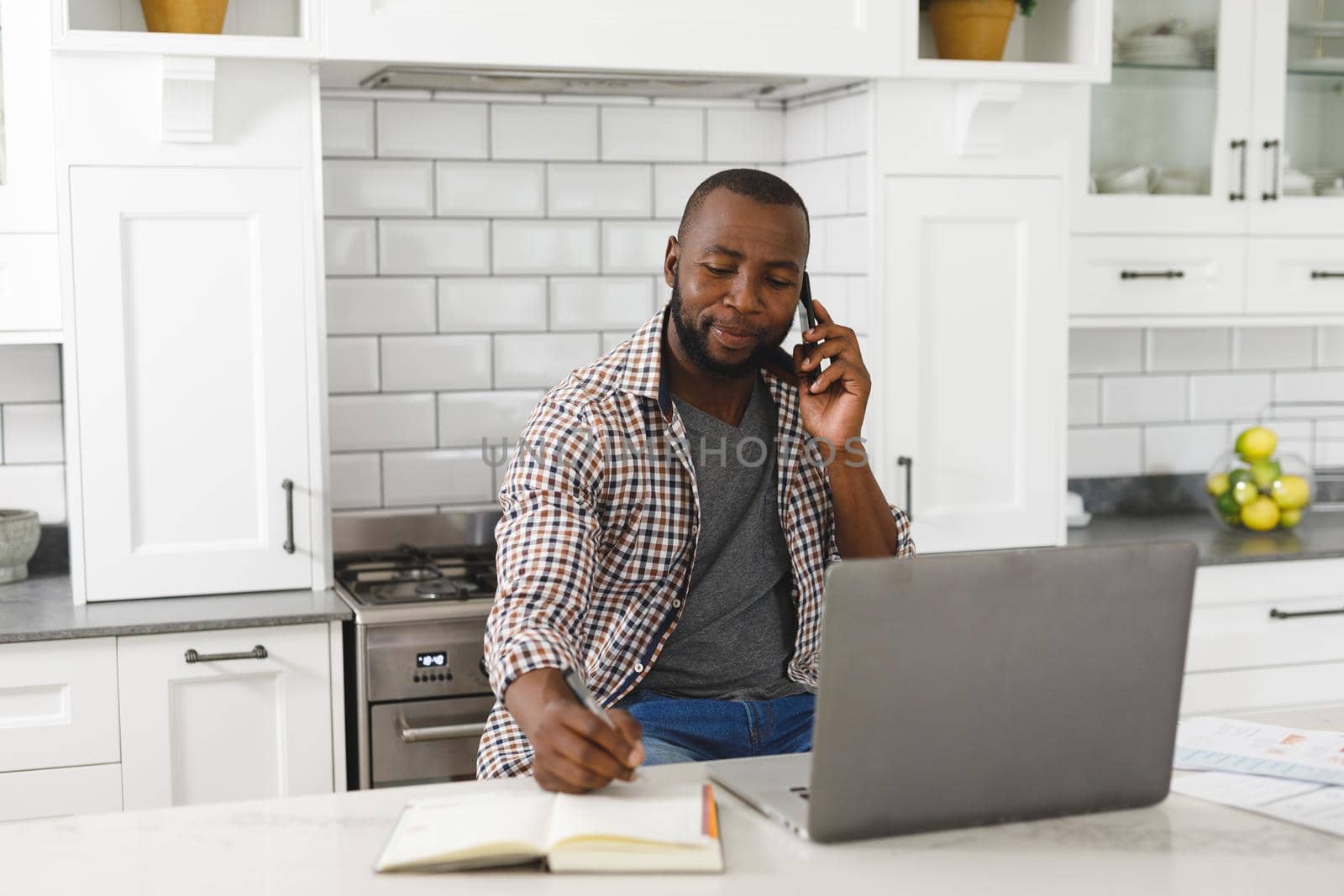 African american man sitting in kitchen working using laptop and smartphone. remote working from home with technology.