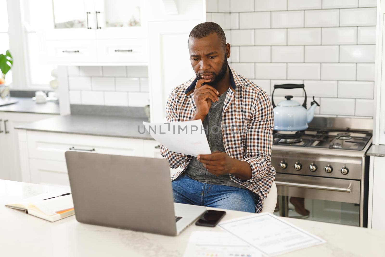 Serious african american man sitting in kitchen working looking at paperwork and using laptop. remote working from home with technology.