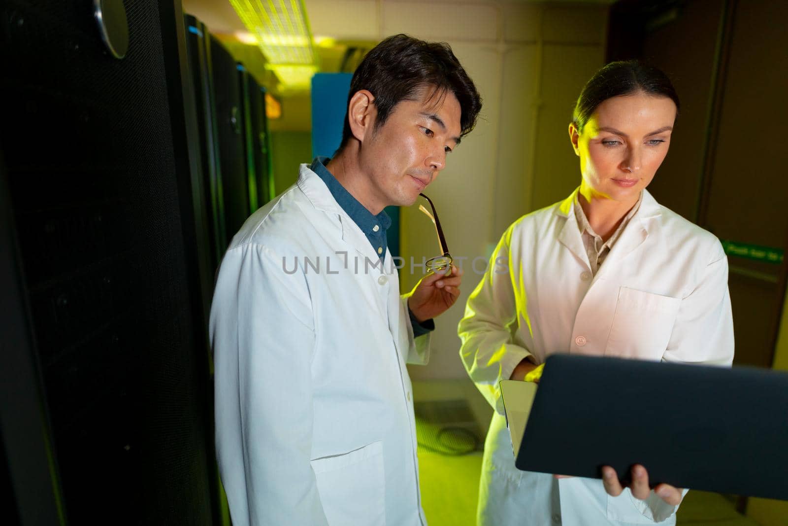 Diverse male and female engineers wearing aprons using laptop while inspecting in server room. database server management and maintenance concept